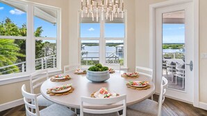 A dining area with a round table, white chairs, and a chandelier. There is a view of a body of water and greenery through large windows. - 400 Steps to Sand Waterfront Luxury w Dock Pool (St. Augustine)