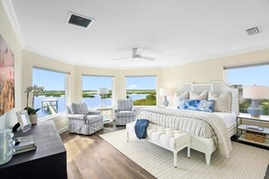 Une chambre spacieuse avec un grand lit, deux fauteuils, un banc et une vue sur un lac, grâce à de grandes fenêtres.