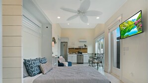 A bedroom with a bed, a ceiling fan, a TV, and a kitchen area in the background. - 100 Steps to Beach Deck w Ocean View Beach Gear (St. Augustine)