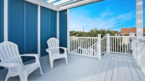 A covered balcony with two white Adirondack chairs, a white railing, and a view of the ocean and buildings in the background. - 100 Steps to Beach Deck w Ocean View Beach Gear (St. Augustine)