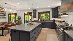 A modern kitchen with a large island, dark cabinetry, and a gas stove. There are pendant lights, a dining area with a long table, and large windows offering a view of greenery.