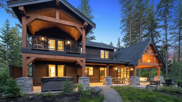 A two-story log cabin with a balcony, stone pillars, and a patio area with outdoor seating.
