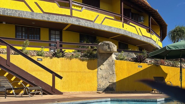 A pool area with a yellow building, stone wall, and a stone pillar. - Pousada Dos Atobás (Arraial do Cabo)