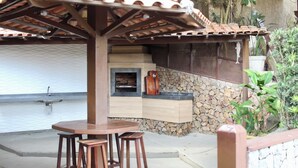 An outdoor kitchen area with a stone wall, a grill, and a wooden table with stools. - Pousada Dos Atobás (Arraial do Cabo)