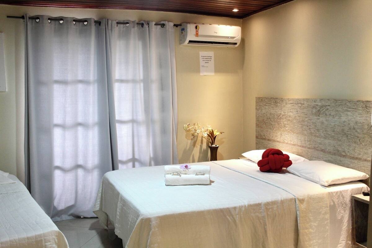 A hotel room with two beds, a large window with curtains, an air conditioning unit, a vase with flowers, and a red pillow.