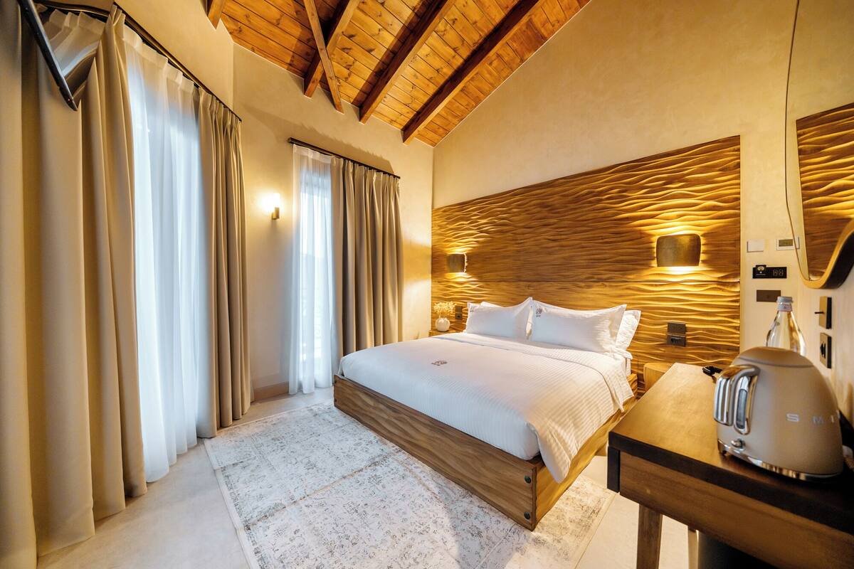 A hotel room with a wooden ceiling, a bed with white linens, a nightstand with a kettle, and a wall-mounted light fixture.