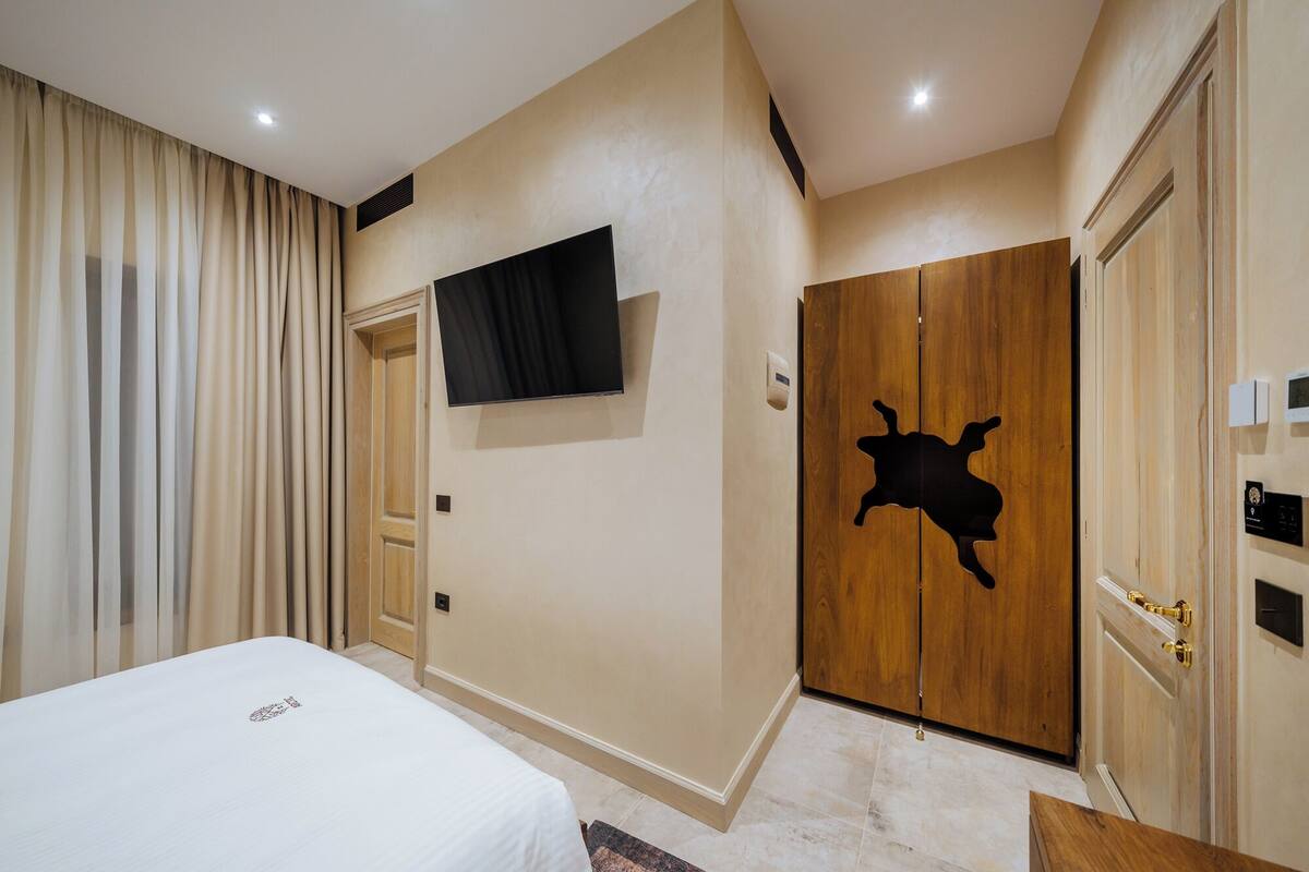 A hotel room with a bed, a TV mounted on the wall, a wooden wardrobe with a unique design, and a door with a light switch.