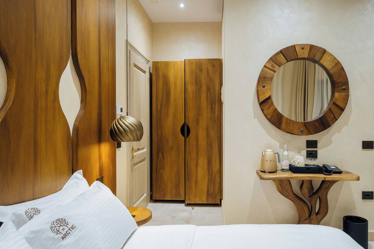 A hotel room with a bed, wooden wardrobe, a round mirror, and a small wooden table with a kettle and a bottle.