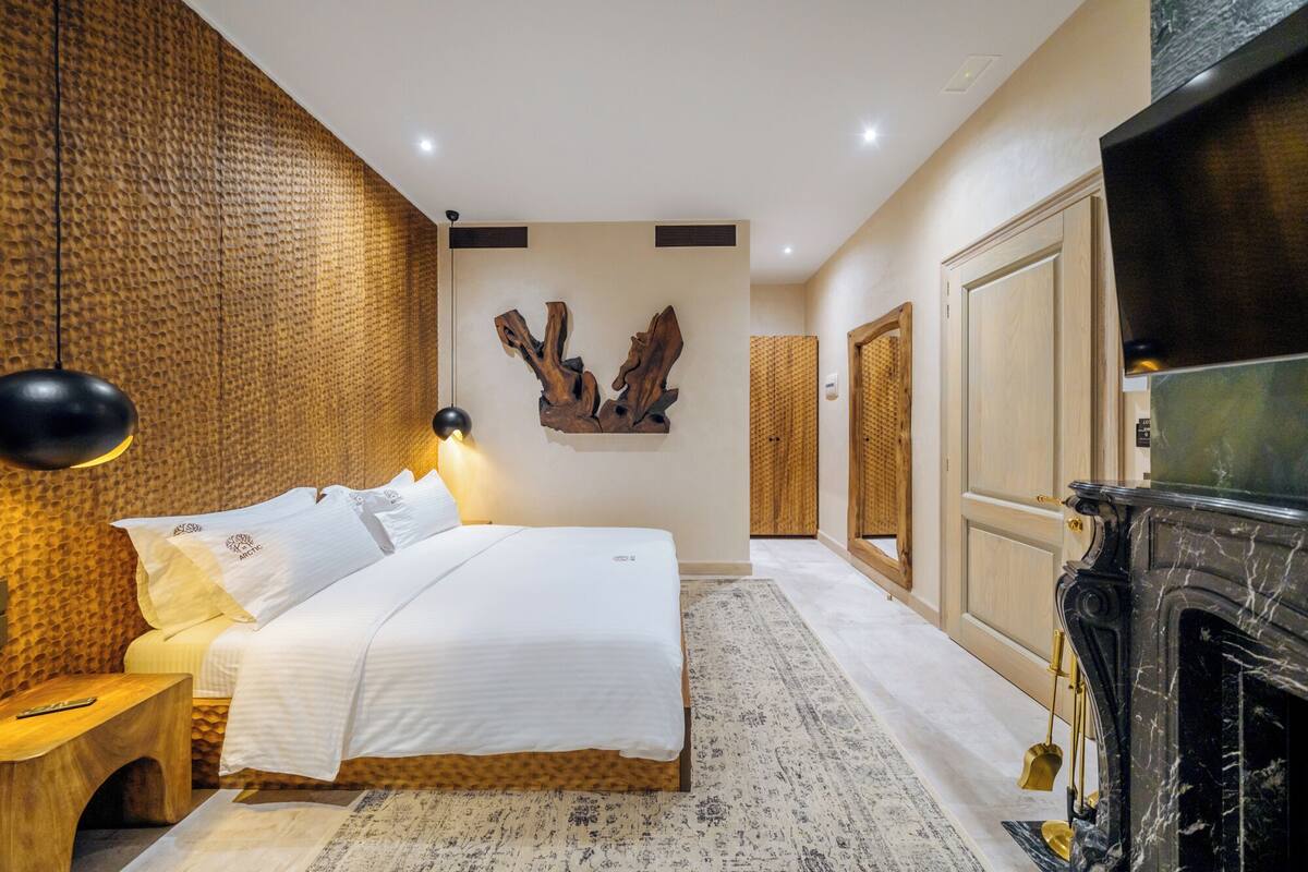 A bedroom with a bed, a wooden headboard, a wall-mounted light, a fireplace, and a mirror.