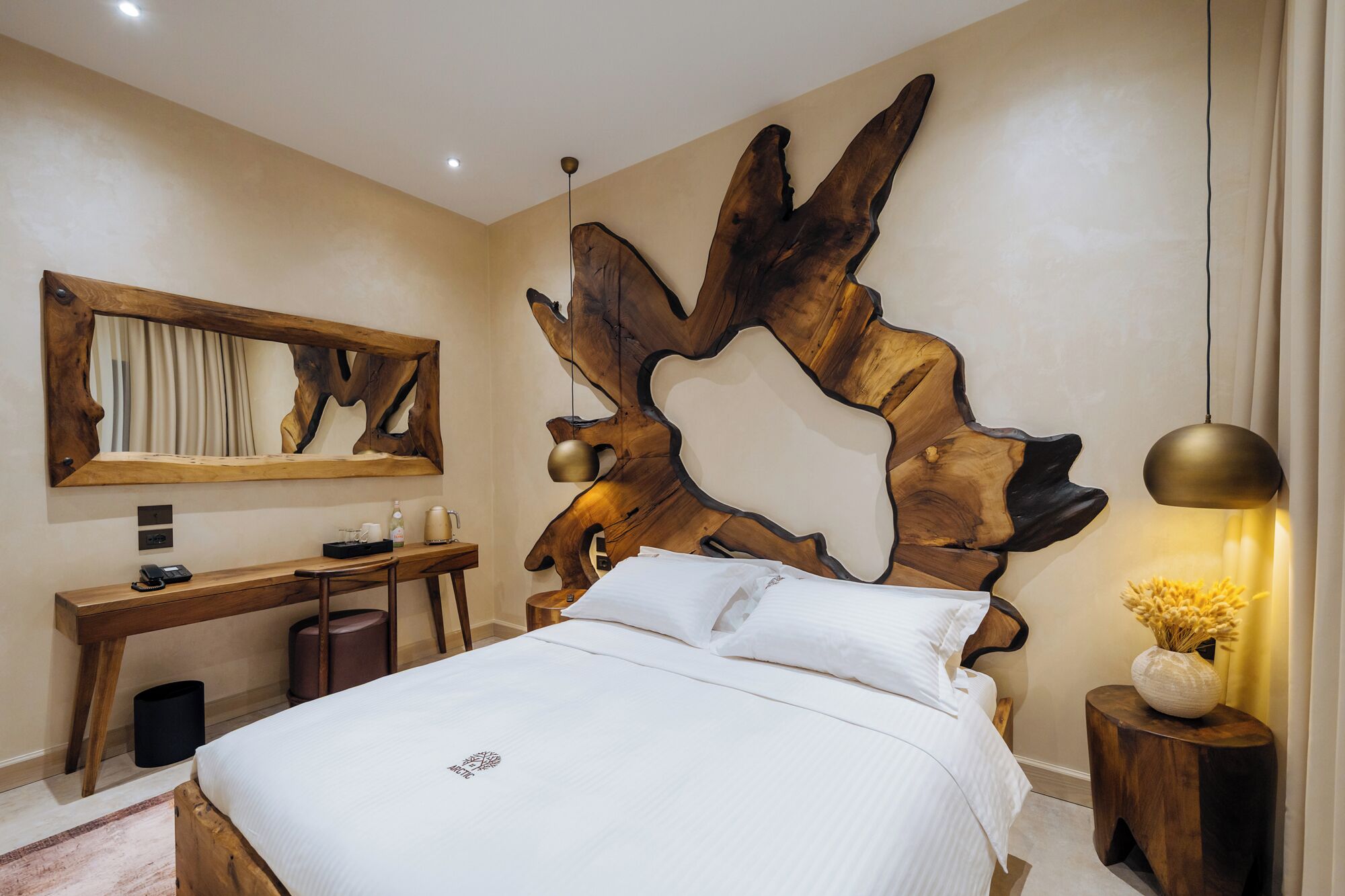 Habitación de hotel con un gran cabecero de madera, un escritorio con silla, cama con ropa blanca y mesita de noche con lámpara y florero.