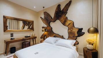 Habitación de hotel con un gran cabecero de madera, un escritorio con silla, cama con ropa blanca y mesita de noche con lámpara y florero.
