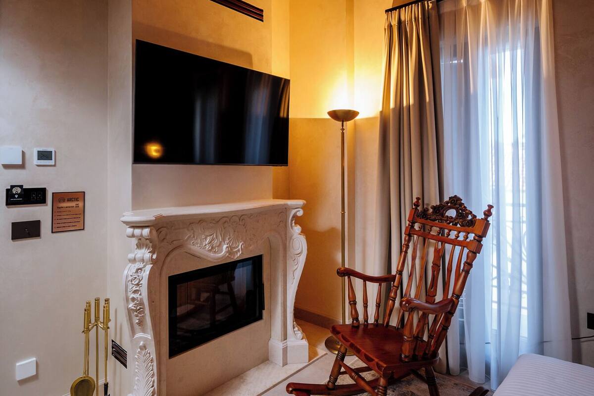 A bedroom with a fireplace, a rocking chair, a bed, a television, and a window with curtains.
