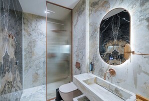 Un baño moderno con pared de mármol, cabina de ducha de vidrio, espejo redondo y lavamanos con grifo de cobre.
