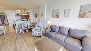 A living room with a grey sofa, a patterned armchair, a wooden coffee table, and a dining area with white chairs and a kitchen in the background. - Master on Gulf & close to Everything & Free round of Golf & Activities Daily! (Panama City Beach)