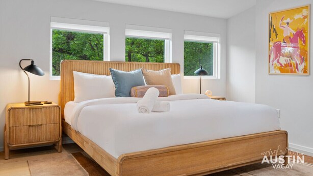 A modern bedroom with a wooden bed frame, white bedding, and three windows offering a view of greenery.