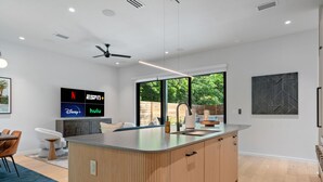 A modern kitchen with a central island, a dining area with chairs, a TV displaying streaming services, and a large window with a view of greenery.