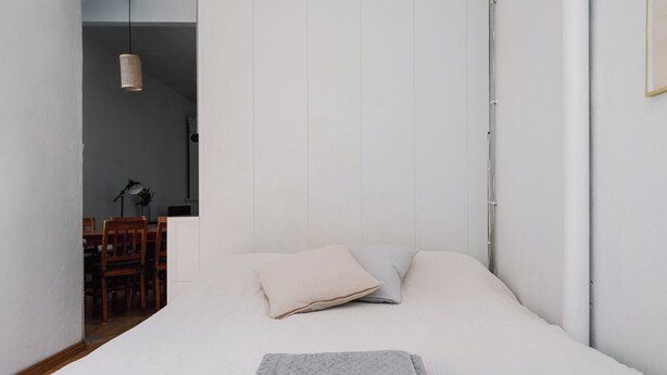 A minimalist bedroom with a neatly made bed, a dining area in the background, and a hanging light fixture. - Vintage 1BR Apt in Kamppi  (Helsinki)