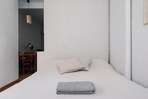 Un dormitorio minimalista con una cama bien tendida, una zona de comedor al fondo y una lámpara colgante.