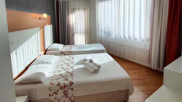 A hotel room with two beds, a wooden floor, a headboard with a light fixture, and large windows with curtains. - Uğur Otel (Kas)