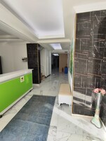 Vestíbulo de hotel con mostrador de recepción, un mostrador verde, alfombra azul, banca blanca y pared de mármol negro con extintor.