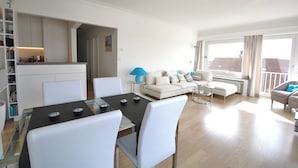 A modern living room with a dining area, white furniture, and a view through large windows. - Apartment in Knokke Near the Beach (Knokke-Heist)