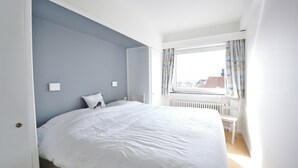 A bedroom with a large bed, a window with curtains, and a radiator. - Apartment in Knokke Near the Beach (Knokke-Heist)
