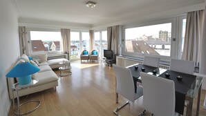 A spacious living area with a sofa, chairs, a dining table, and large windows offering a city view. - Apartment in Knokke Near the Beach (Knokke-Heist)