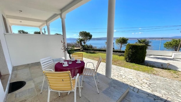 Een overdekt terras met een tafel en stoelen, uitzicht op een waterpartij, en een pad van stenen.