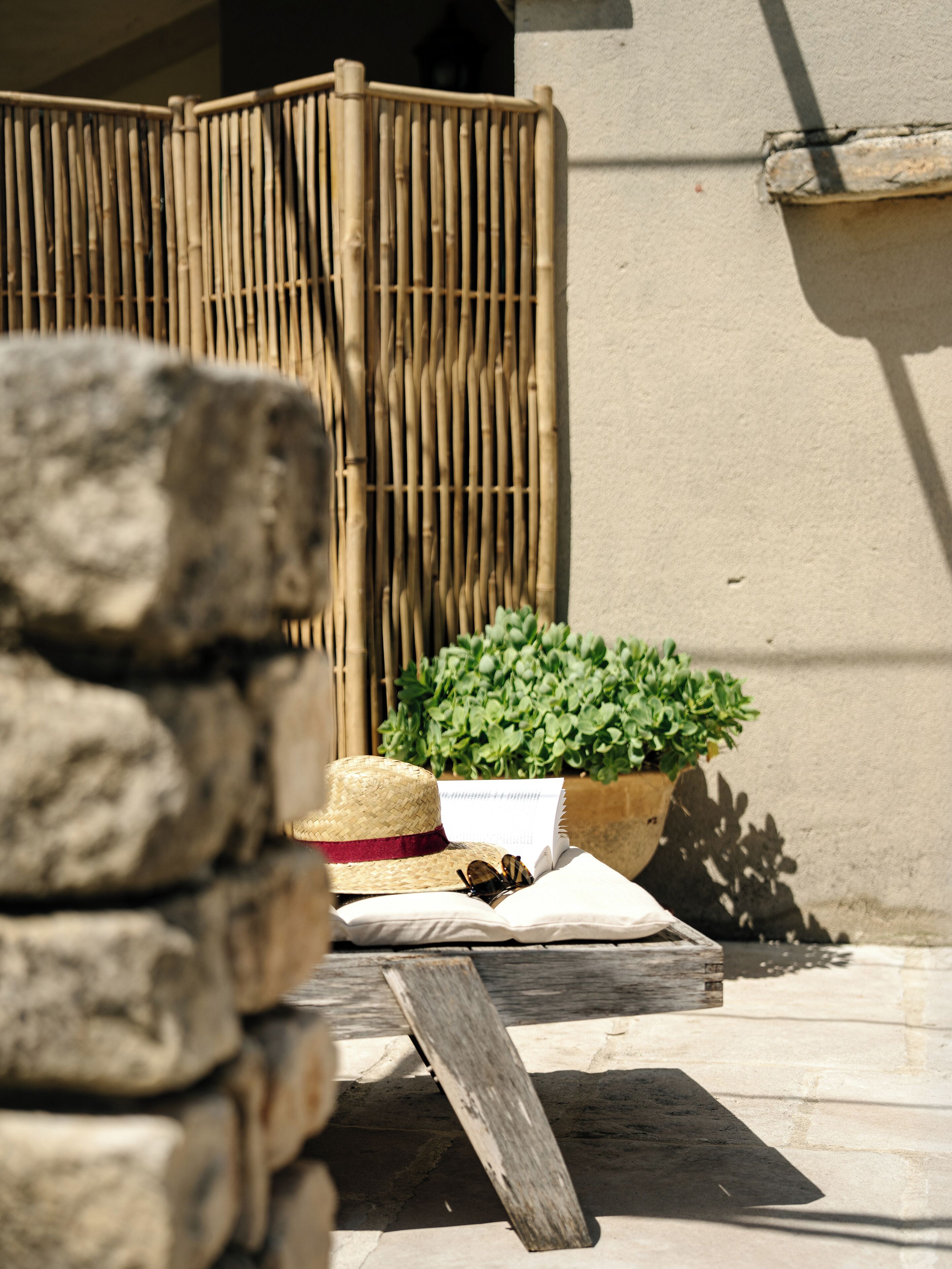 A stack of stones, a straw hat, a book, and a potted plant on a wooden table outside a building with a bamboo fence and a concrete wall.