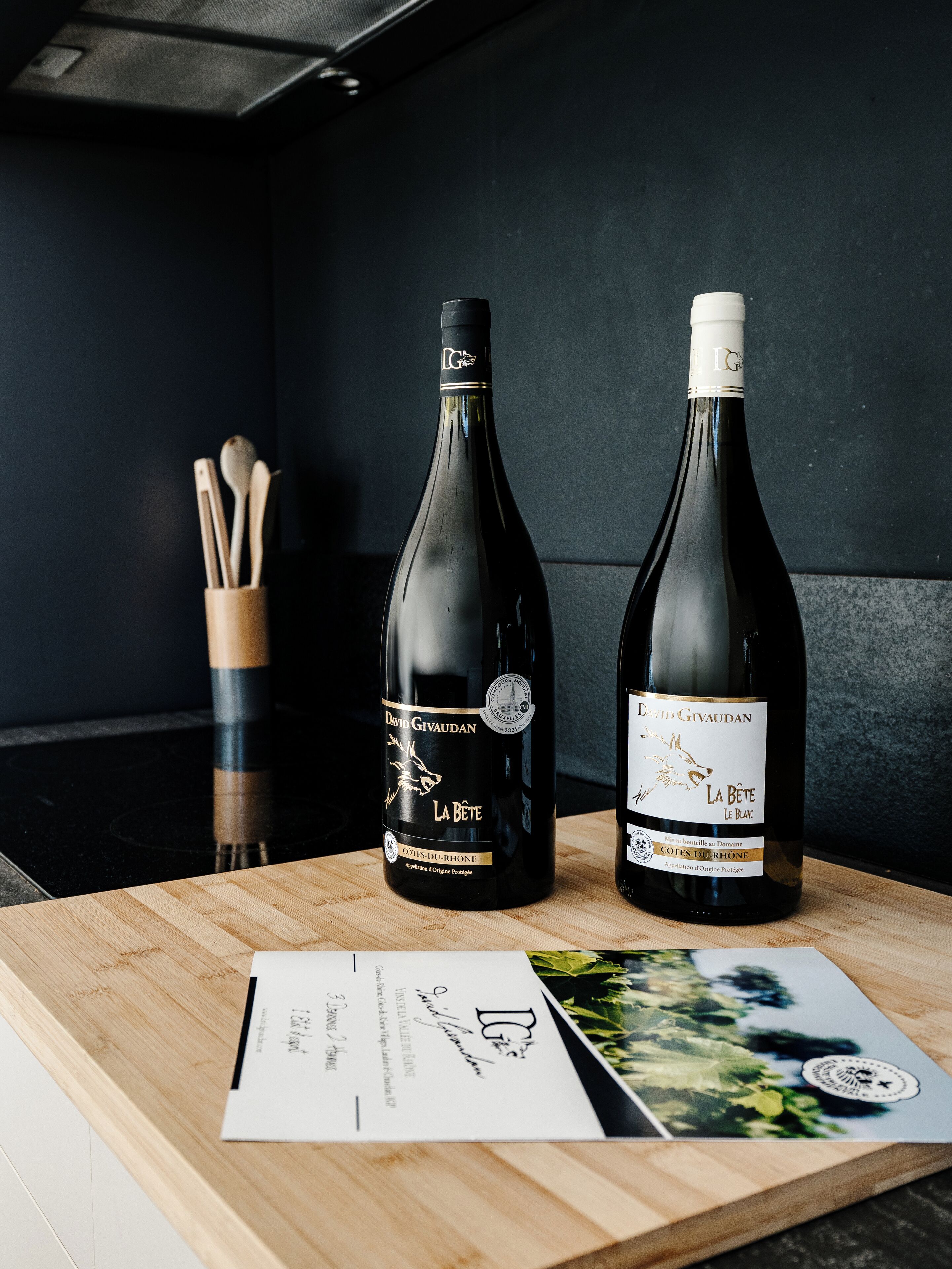 Two wine bottles on a wooden table with a menu and a spoon holder in the background.