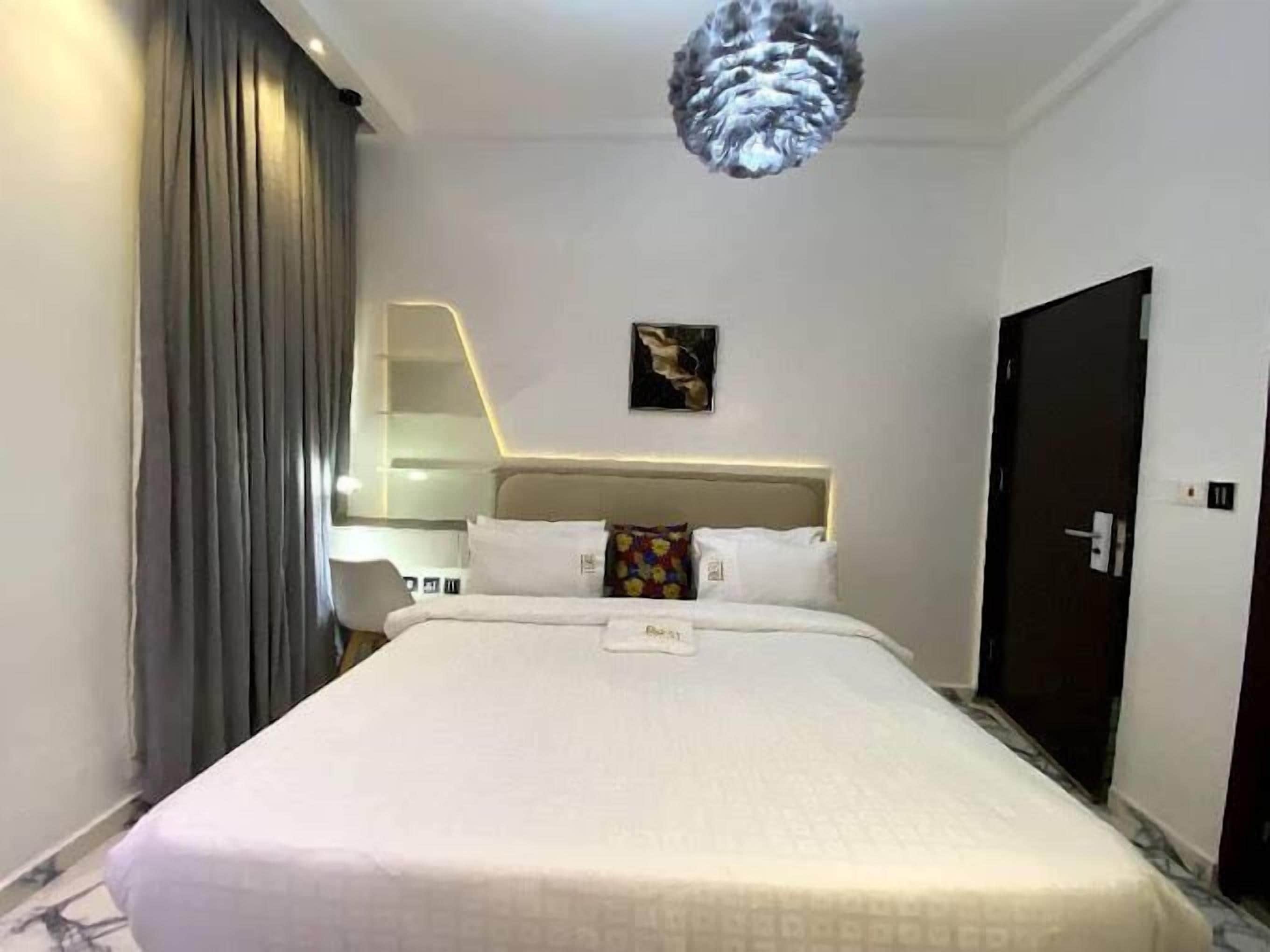 A hotel room with a large bed, a headboard with integrated lighting, a painting on the wall, and a door to the right.