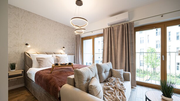 Ein modernes Schlafzimmer mit einem großen Bett, einer Couch, einem kleinen Tisch mit einer Pflanze und Blick auf die Stadt durch große Fenster.