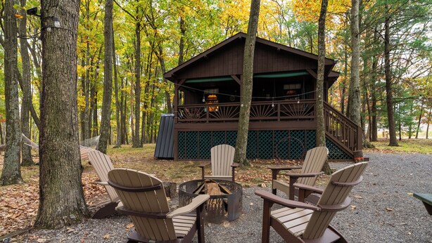 Una cabaña con terraza, rodeada de árboles, amueblada con sillas Adirondack y un brasero.