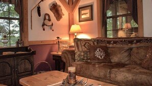 Una sala de estar acogedora con una mesa de madera, un sofá marrón con cojines estampados, una lámpara de pared y decoración rústica que incluye calaveras de animales y un cuadro enmarcado.