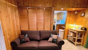 A cozy room with a brown sofa, wooden blinds, a kitchen area, and a small wooden shelf.