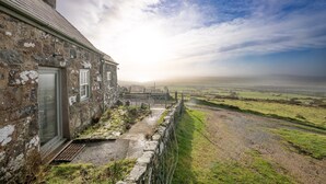 A stone house with a door and windows, a stone wall, and a grassy field in the background. - 2-Bryn-Tirion | Charming Traditional Cottage with Stunning Sea Views (Pwllheli)