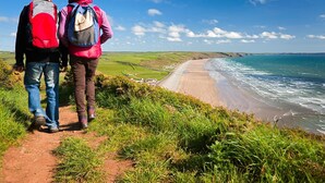 Two people walking on a dirt path beside a beach with a clear view of the ocean and distant hills. - 2-Bryn-Tirion | Charming Traditional Cottage with Stunning Sea Views (Pwllheli)