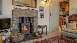 A cozy living room with a stone fireplace, a television, and a comfortable armchair. - 2-Bryn-Tirion | Charming Traditional Cottage with Stunning Sea Views (Pwllheli)