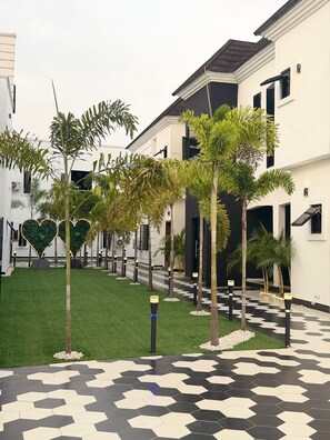 A courtyard with a geometric tile floor, palm trees, and a building with a dark roof and white walls. - Emiez Apartment and Hotel  (Benin City)