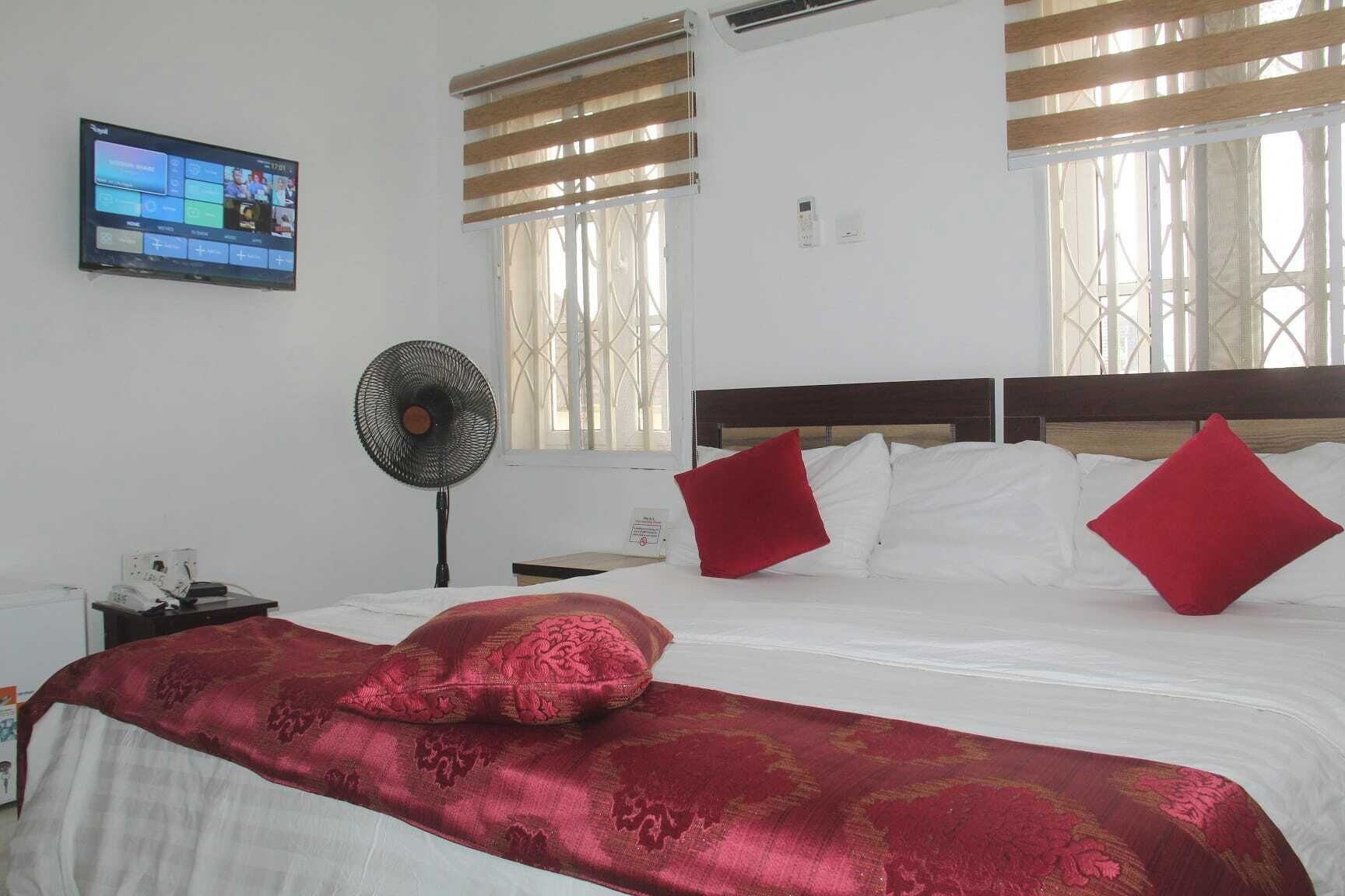 Habitación de hotel con cama, televisor montado en la pared, ventilador y ventana con persianas.