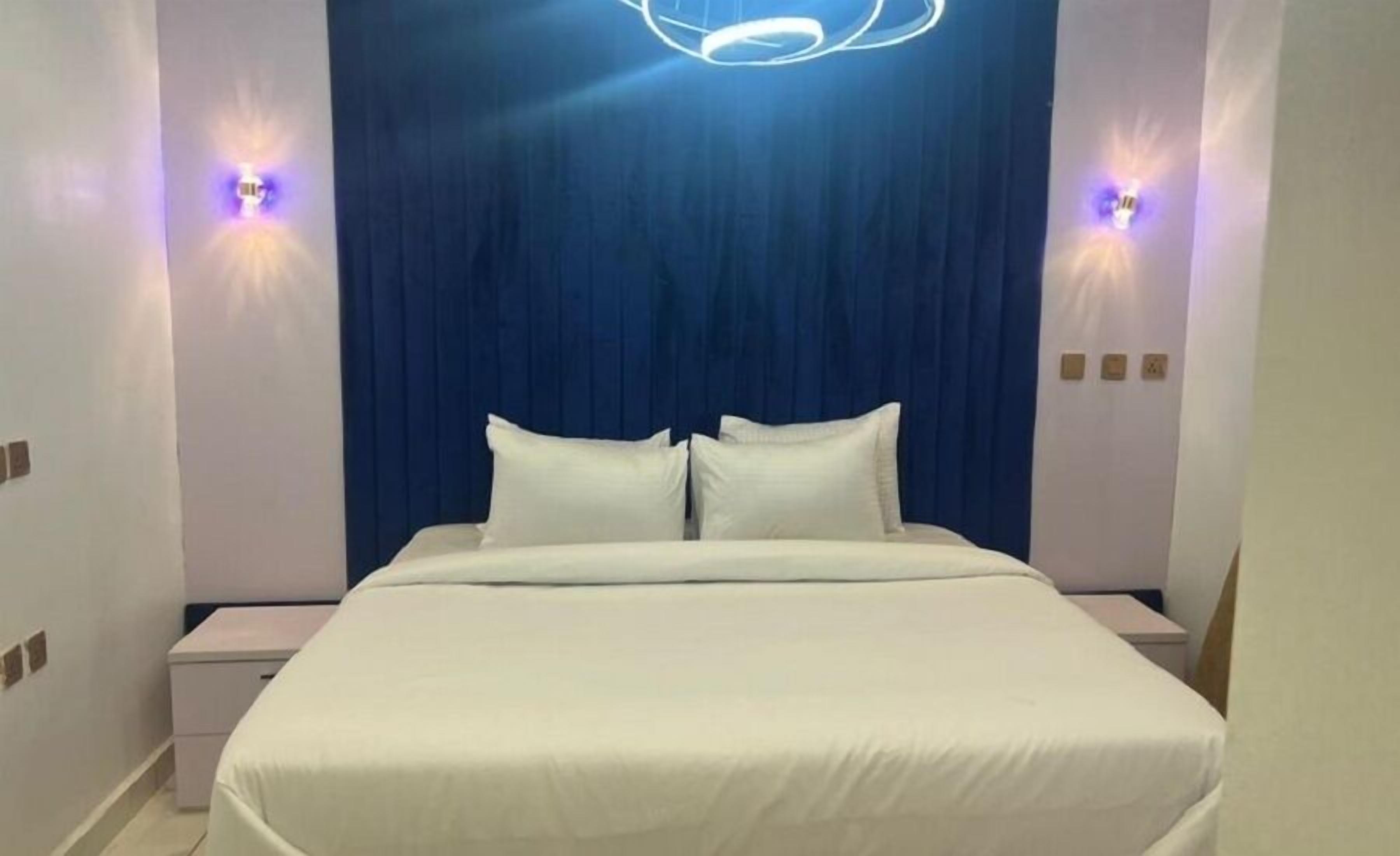 A modern hotel room with a large bed, two bedside tables, and a blue curtain backdrop.