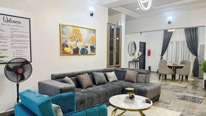 A modern living room with a sectional sofa, a round coffee table, and a fan. There is a dining area with a table and chairs in the background.