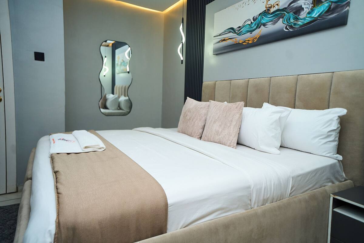 A neatly made bed with a beige headboard, a wall-mounted mirror, and a painting.
