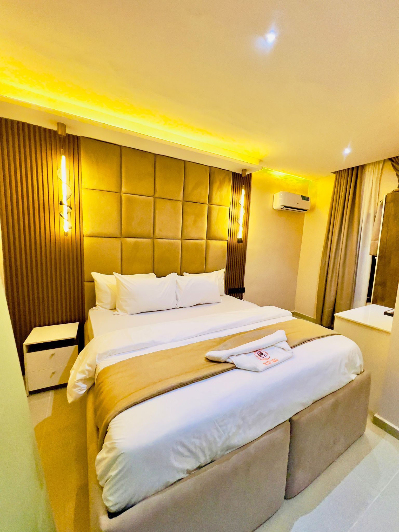 A hotel room with a large bed, a bedside table, an air conditioning unit, and a mirror.