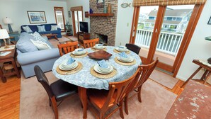 Un comedor con una mesa redonda dispuesta para seis personas, rodeada de sillas de madera. Se observa una chimenea de ladrillo al fondo, y la habitación tiene un balcón con vistas a casas vecinas.