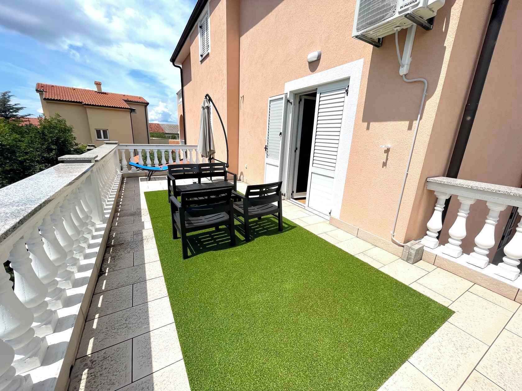 Un balcón con mesa y sillas, barandal blanco y zona de césped artificial verde.