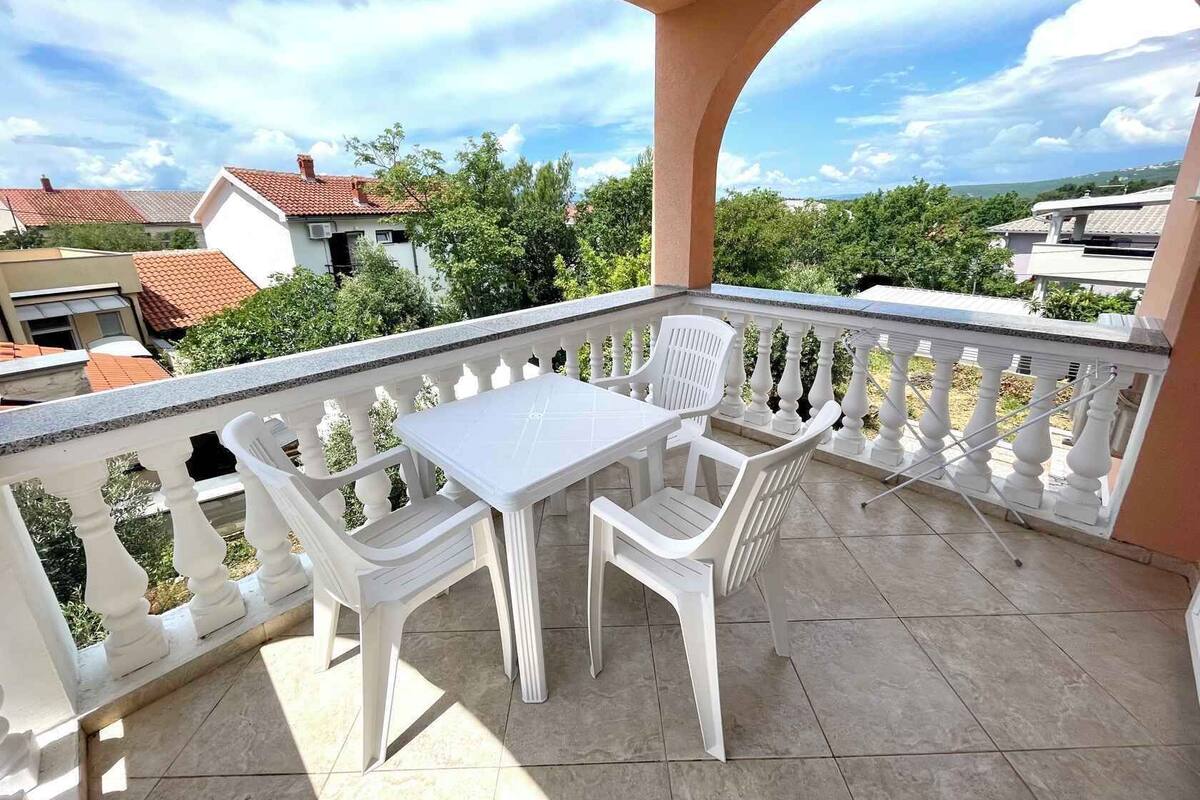 A balcony with a white table and chairs, a tiled floor, and a view of surrounding buildings and greenery.