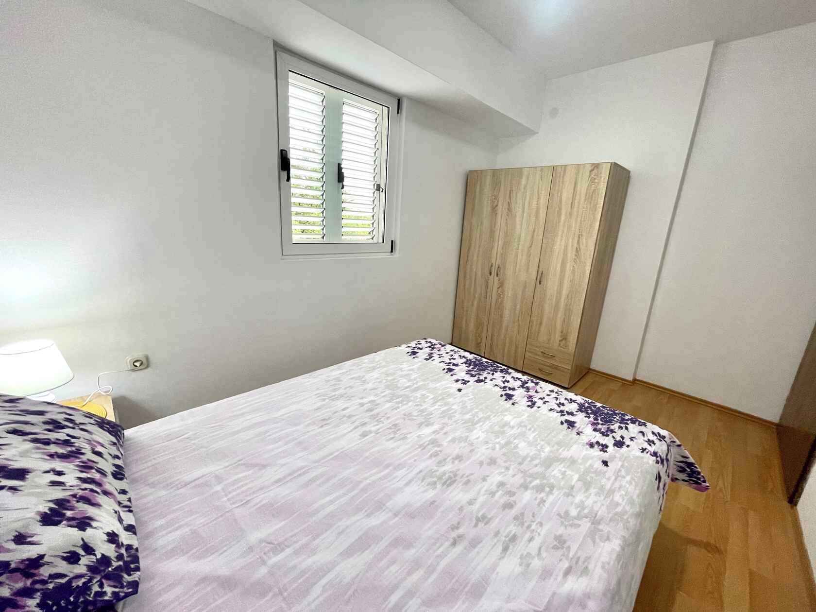 A bedroom with a bed, a window with blinds, a wardrobe, and a nightstand with a lamp.