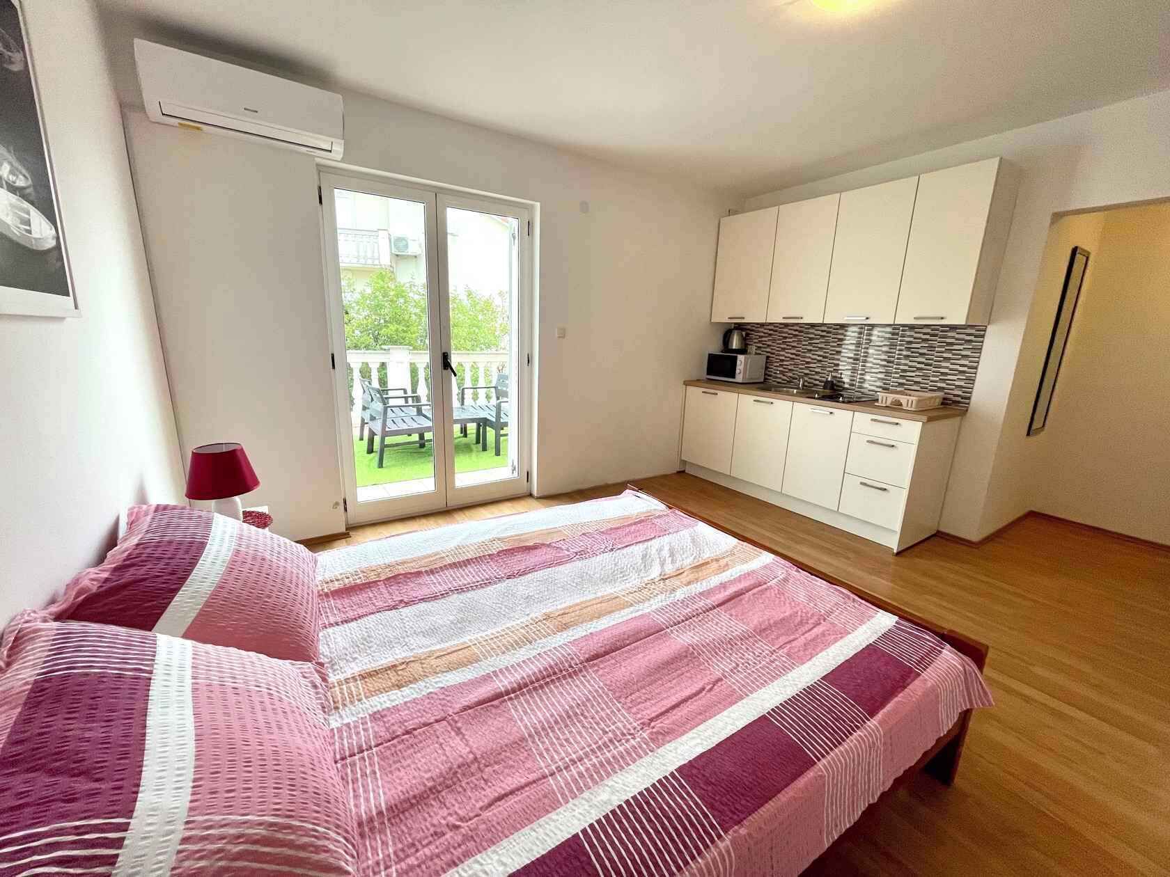 A bedroom with a bed, a sliding glass door to a balcony, a kitchenette, and a small table with a lamp.