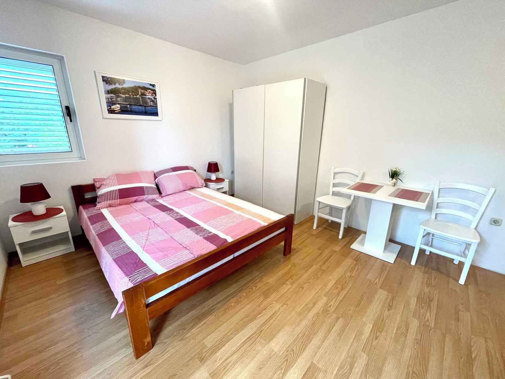A bedroom with a bed, bedside tables, a wardrobe, a desk with chairs, and a window with blinds.